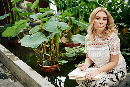 Beautiful serious young woman spending time in flower garden and drawing plants