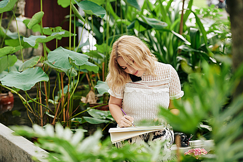 Creative young woman sitting in flower garden, talking on phone with client and drawing sketch