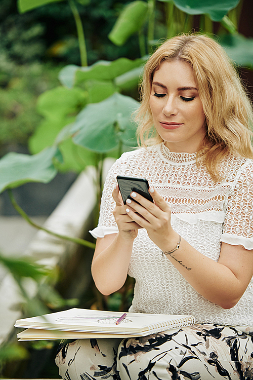 Portrait of young beautiful Caucasian woman checking application on smartphone when sitting outdoors