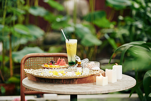 Tall glass of delicious cocktail and plate of cut fruits on small table in backyard on spa hotel