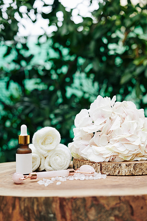 Bottle of moisturizing facial serum and quartz massage roll on wooden stump with blooming flower and small rolled towels