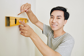 Cheerful young Asian man drawing line when measuring wall in his new apartment