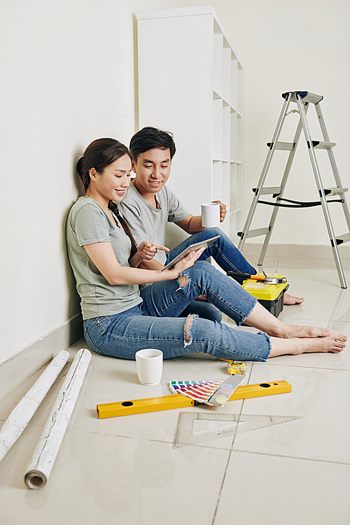 Cheerful young Asian couple having coffee break and looking at inspiring photos on tablet computer after assembling storage unit in new apartment