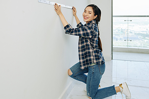 Beautiful young Vietnamese woman in jeans and plaid shirt measuring walls with metal ruller