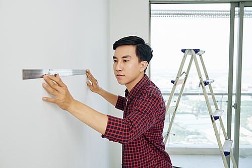 Serious young Asian man measuring walls in his new house with metal ruller before start painting