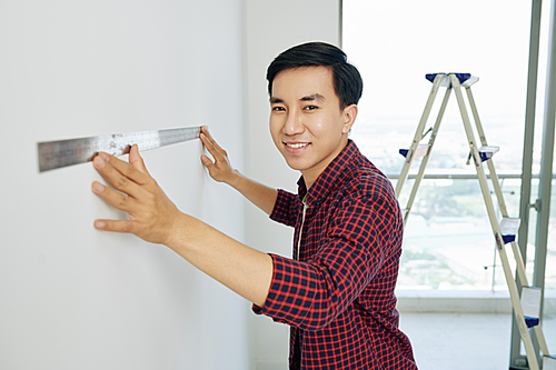 Handsome cheerful Vietnamese man in plaid shirt using metal ruller when preparing for house renovation