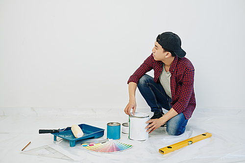 Pensive young man in cap looking at blank white wall in his new apartment when opening can of paint