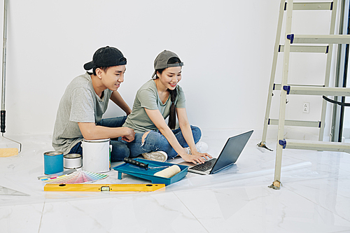 Cheerful young Asian couple in caps sitting on the floor in new apartment with tools and paints around and seaching for design ideas online