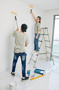 Cheerful young Vietnamese couple in jeans and t-shirts painting room walls with white paint