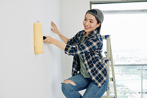 Happy young Asian woman in cap and plaid shirt priming wall before applying plaster