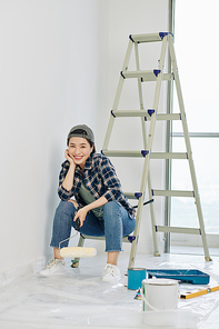 Happy young Vietnamese woman resting on ladder with roller in hand after painting walls in room