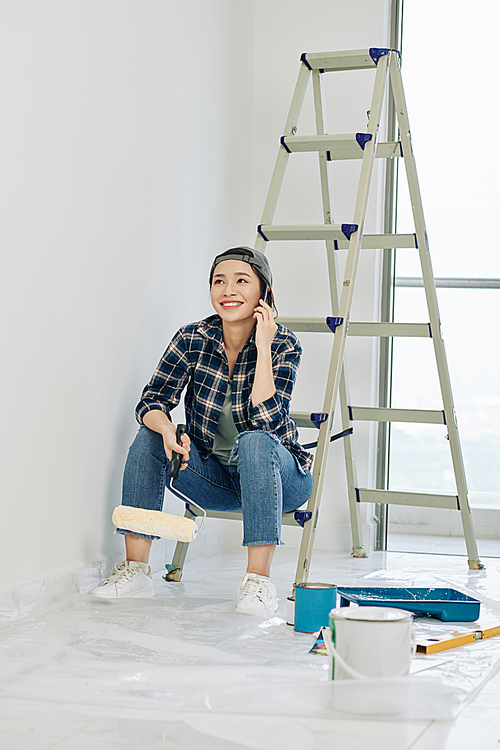 Cheerful young Vietnamese woman sitting on ladder and calling on phone after painting rooms in her new apartment