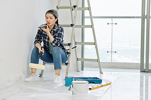 Frowning young Vietnamese woman recording voice message to friend when having short break after painting walls in house