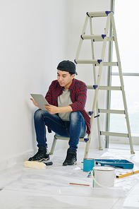 Handsome young Vietnamese man sitting of ladder and reading instruction how to paint walls with roller