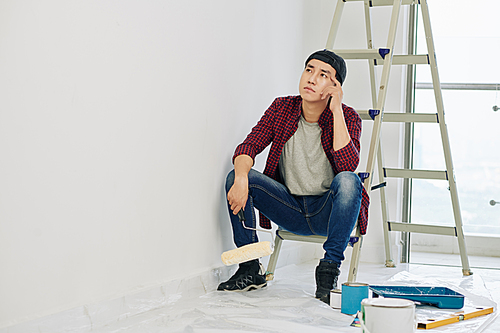 Pensive tired young Vietnamese man with roller in hand sitting on ladder and looking at wall he just painted