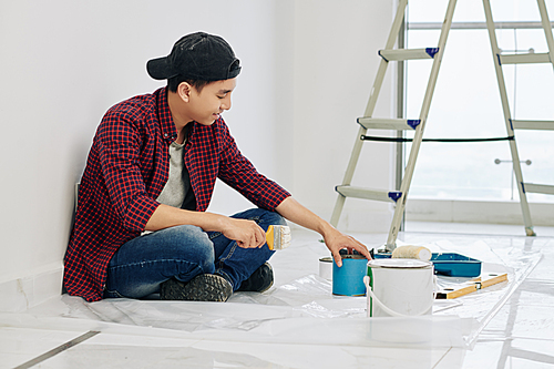 Smiling young Asian man in cap and plaid shirt opening can of paints to mix them before painting walls in apartment