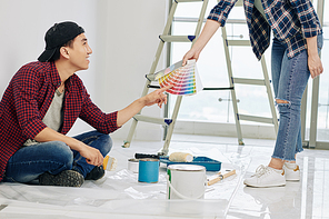 Smiling young Asian man pointing at color in palette of swatches in hand of girlfriend and choosing shade for bedroom walls