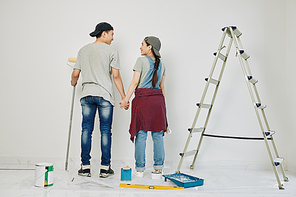 Young smiling Asian couple holding hands and looking at each other before starting painting wall in their new house