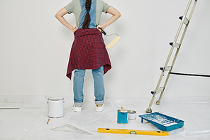 Young proud woman standing with hands on hips and looking at wall she just painted, view from the back