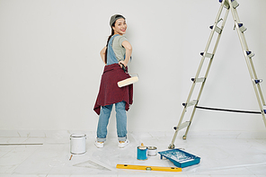 Cheeful young Vietnamese woman with roller in hand turning back and  after painting wall