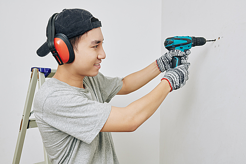 Smiling young Vietnamese man wearing protective ear muffs and gloves when drilling wall in apartment