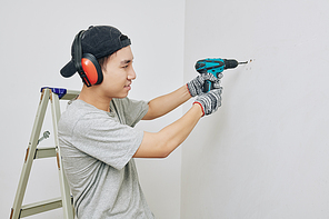 Young Asian man in protective ear muffs drilling hole in wall for hanging pictures