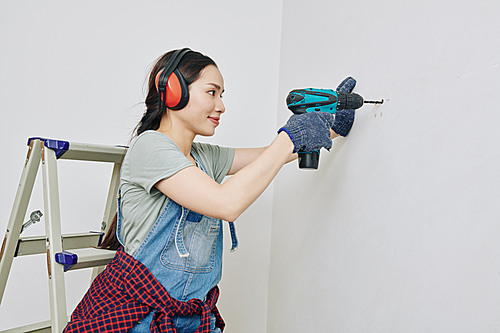 Smiling pretty young woman in protective ear muffs drilling wall in her apartment