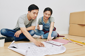 Cheerful young Asian couple sitting on the floor and discussing construction plan