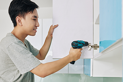 Handsome young Vietnamese man using electric screwdriver when assembling cupboard in kitchen