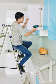 Young Asian man sitting on ladder when fixing door of kitchen cupboard