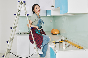 Smiling attractive young Vietnamese woman sitting on ladder with screwdriver in hand in kitchen where she is assembling cupboards