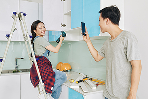 Young Asian man with smartphone filming his pretty girlfriend fixing door of kitchen cupboard for his diy home renovation blog