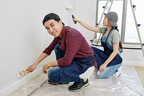 Cheerful young Asian man painting wall with small brush when his girlfriend using foam roller
