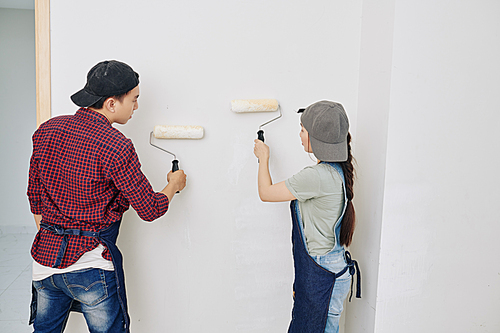 Young Asian people applying coat of matte white paint on room walls in new apartment