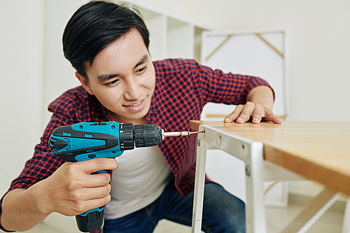 Positive young Vietnamese man assembling furniture in new apartment and tightening screw with electric screwdriver