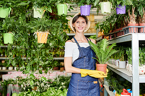 Portrait of pretty female greenhouse worker in blue apron and rubber gloves holding pot with plant