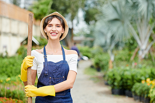 Portrait of pretty smiling woman in overall and hat standing with spade or rake wooden handle in garden