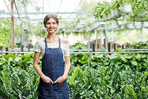 Positive pretty young woman standing in front of greenhouse full of various plants and flowers and smiling at camera