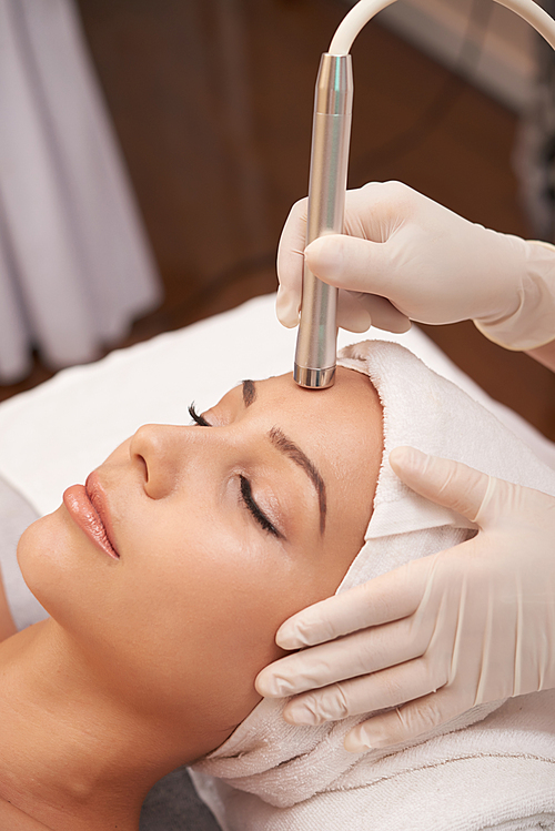Close-up of Asian beautiful woman with towel on her head relaxing while cosmetologist making ultrasound facial procedure
