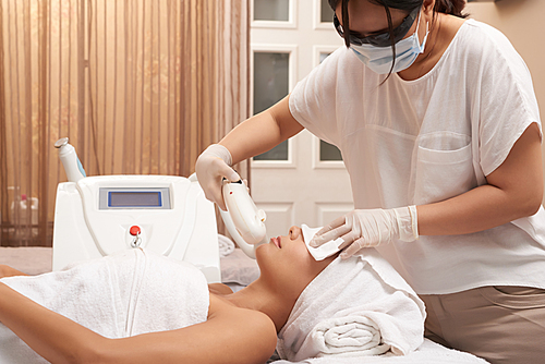 Beautician in protective mask and gloves making ultrasound procedure with special equipment for woman's face while she lying and relaxing in spa salon