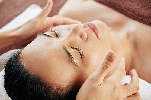 Relaxed young Asian woman enjoying relaxing anti-aging face massage in beauty salon
