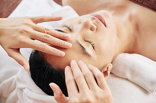 Hands of beautician massaging forehead of young woman relaxing on massage table