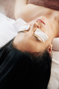 Pretty young Asian woman lying on massage bed with textile pads soaked in moisturizing serum on eyelids