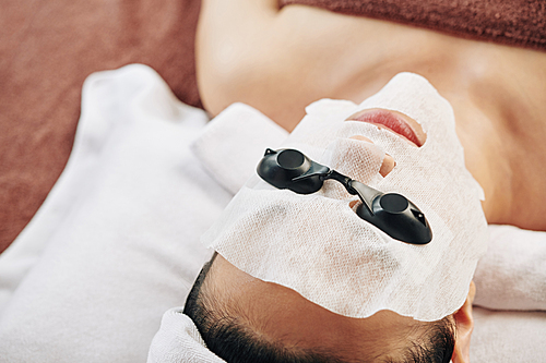 Young woman lying on bed in spa salon with rejuvenating sheet mask and protective glasses ready for UV therapy