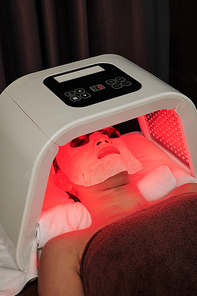 Woman having color light therapy and lying in sheet mask under red light lamp