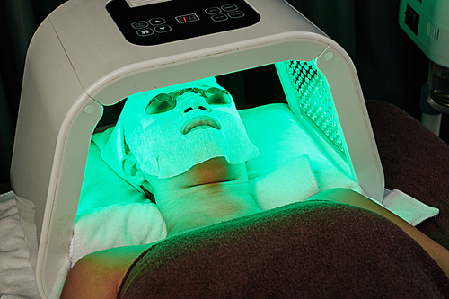 Young woman with sheet mask on her face having LED light treatment in beauty salon
