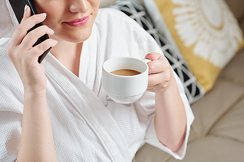 Young woman enjoying cup of delicious morning cappuccino and making phone call when sitting of sofa in bath robe