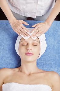 Hands of beautician using cleanser and wiping face of pretty female client with cotton pads, view from above