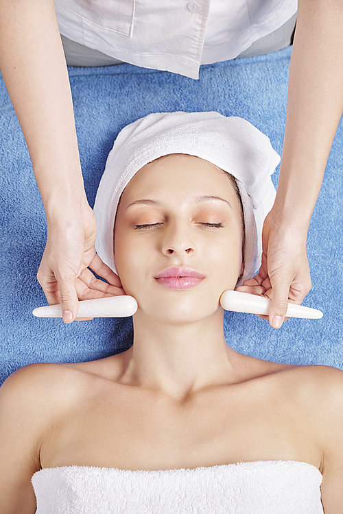 Cosmetologist using stone sticks when massaging face of relaxing young Caucasian woman, view from above