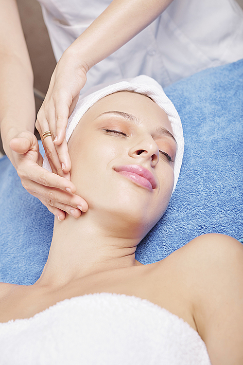 Smiling young Caucasian woman lying on bed and enjoying face lifting massage in beauty salon